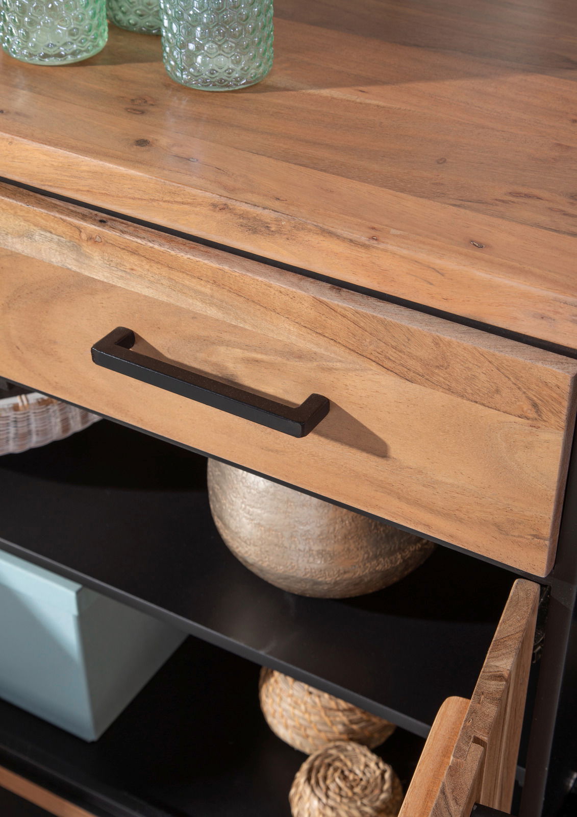 Nahaufnahme eines Sideboards aus Holz mit schwarzem Griff, teilweise geöffnete Schublade und Regalfächer mit Dekorationen, aus der Vogelperspektive.