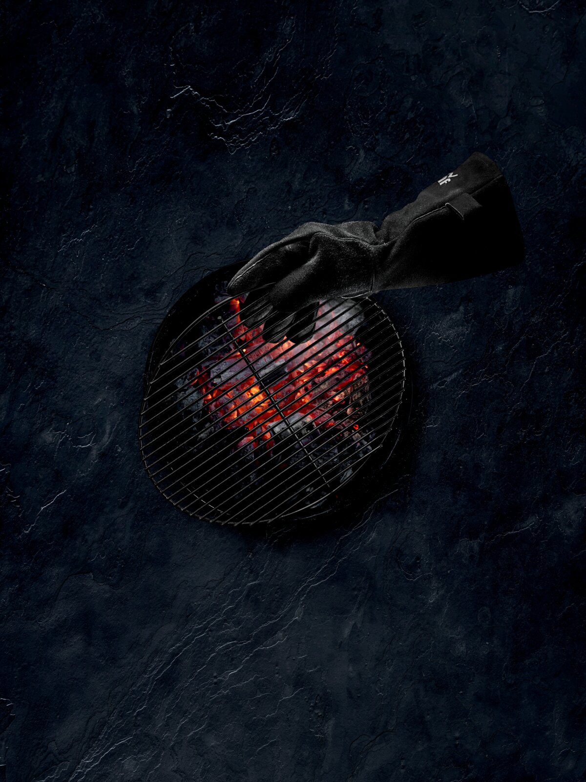 WMF BBQ Grillhandschuhe Een zwarte, hittebestendige barbecuehandschoen wordt van bovenaf in een ronde houtskoolbarbecue met gloeiende kolen gestoken, bovenaanzicht/top-downperspectief