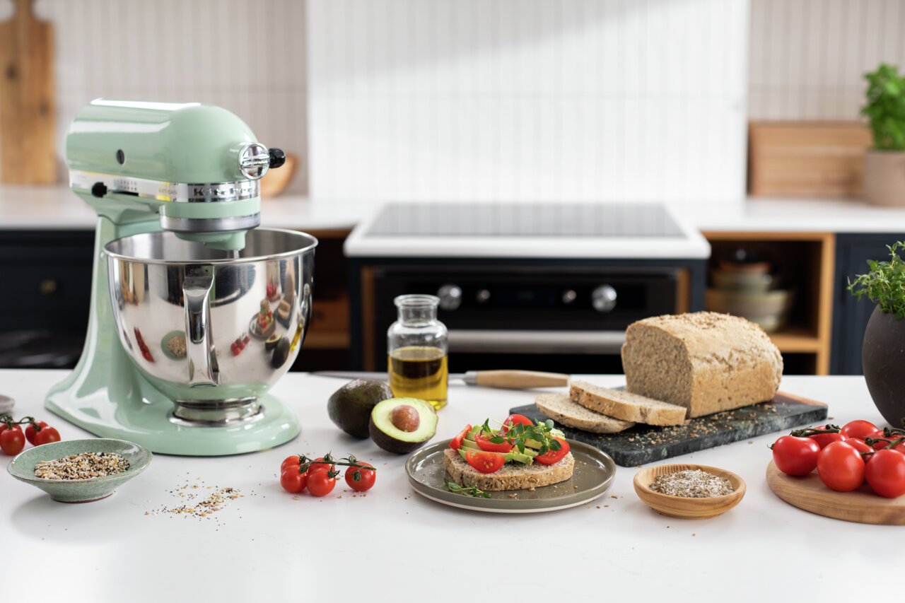 Küchenmaschine Artisan in Pastellgrün auf einer Küchenarbeitsplatte, umgeben von frischen Zutaten wie Tomaten, Avocado und Brot. Perspektive von vorne links.