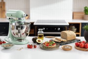 Artisan-keukenmachine in pastelgroen op een keukenblad, omringd door verse ingrediënten zoals tomaten, avocado en brood. Aanzicht van linksvoor.