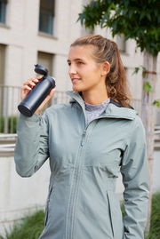 Frau in Outdoor-Jacke hält einen schwarzen Thermobecher mit geöffnetem Deckel in der Hand, seitliche Perspektive