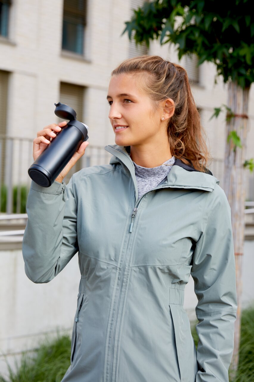 Frau in Outdoor-Jacke hält einen schwarzen Thermobecher mit geöffnetem Deckel in der Hand, seitliche Perspektive