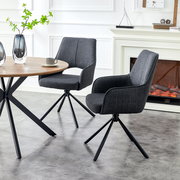 Zwei dunkelgraue, gepolsterte Esszimmerstühle mit Armlehnen und schwarzem Metall-Spiderfuß an einem runden Holztisch, seitliche Perspektive im hellen Wohnraum mit Kamin im Hintergrund.