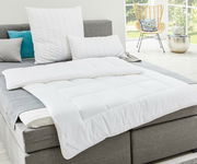 Weiße Faserdecke auf einem grauen Bett, fotografiert aus einer seitlichen Perspektive in einem hellen Raum mit großen Fenstern.