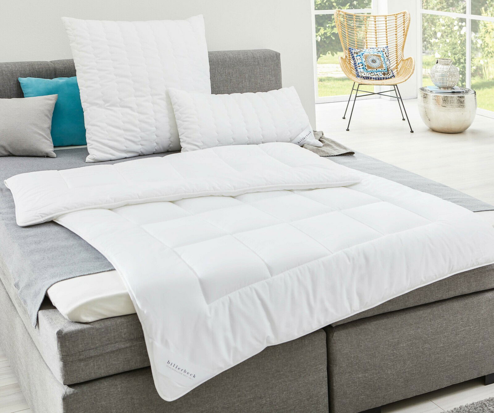 billerbeck Bettdecke UNO ANTIGUA Weiße Faserdecke auf einem grauen Bett, fotografiert aus einer seitlichen Perspektive in einem hellen Raum mit großen Fenstern.