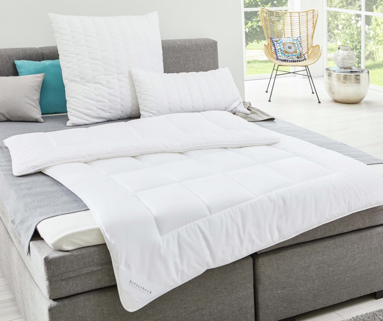 billerbeck Bettdecke UNO ANTIGUA Weiße Faserdecke auf einem grauen Bett, fotografiert aus einer seitlichen Perspektive in einem hellen Raum mit großen Fenstern.