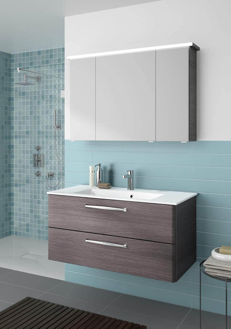 Moderner Badblock mit grauen Schubladen und weißem Waschbecken, frontal betrachtet, vor einer Wand mit blauen Fliesen.