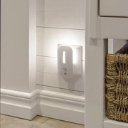 Weißes LED-Nachtlicht mit Bewegungsmelder in der Steckdose, warmes Licht an weißer Holzpaneel-Wand im Flur, seitliche Perspektive auf das Produkt.