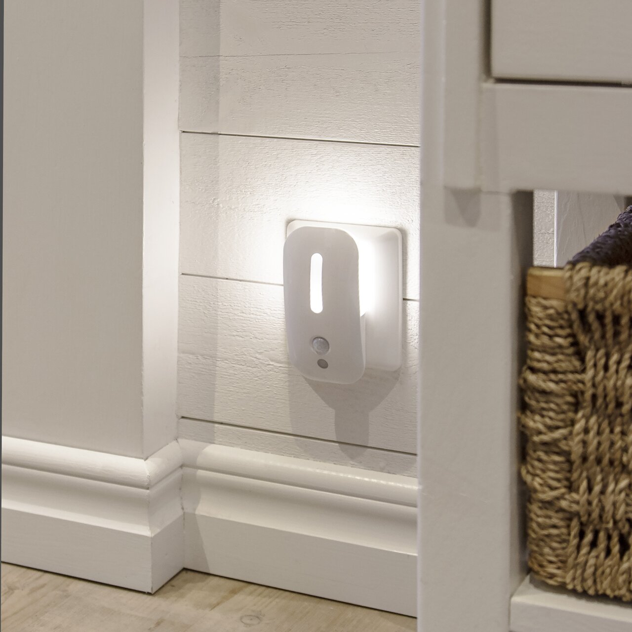 Weißes LED-Nachtlicht mit Bewegungsmelder in der Steckdose, warmes Licht an weißer Holzpaneel-Wand im Flur, seitliche Perspektive auf das Produkt.