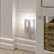 Weißes LED-Nachtlicht mit Bewegungsmelder in der Steckdose, warmes Licht an weißer Holzpaneel-Wand im Flur, seitliche Perspektive auf das Produkt.