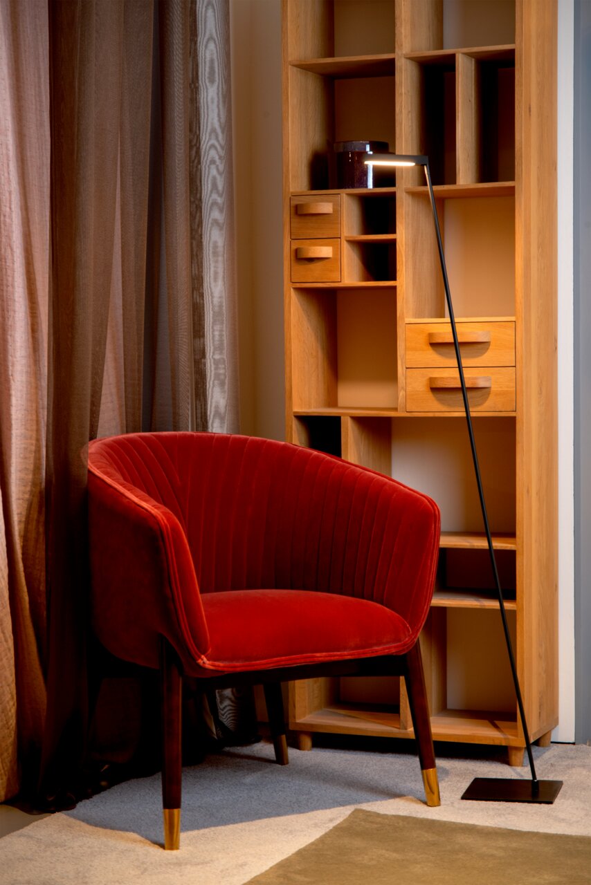 Steh- und Leseleuchte Lucide LAVALE in einem gemütlichen Wohnzimmer, neben einem roten Sessel und einem Holzregal, aus seitlicher Perspektive.