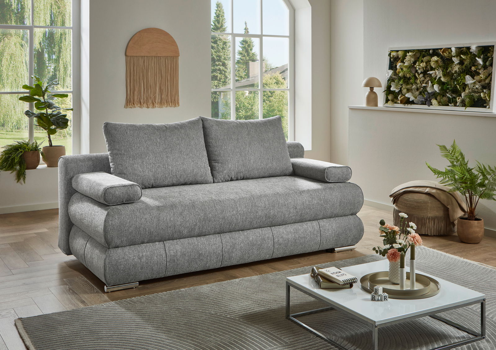 Graues Funktionssofa in einem modernen Wohnzimmer mit großen Fenstern und Pflanzen, seitliche Perspektive.