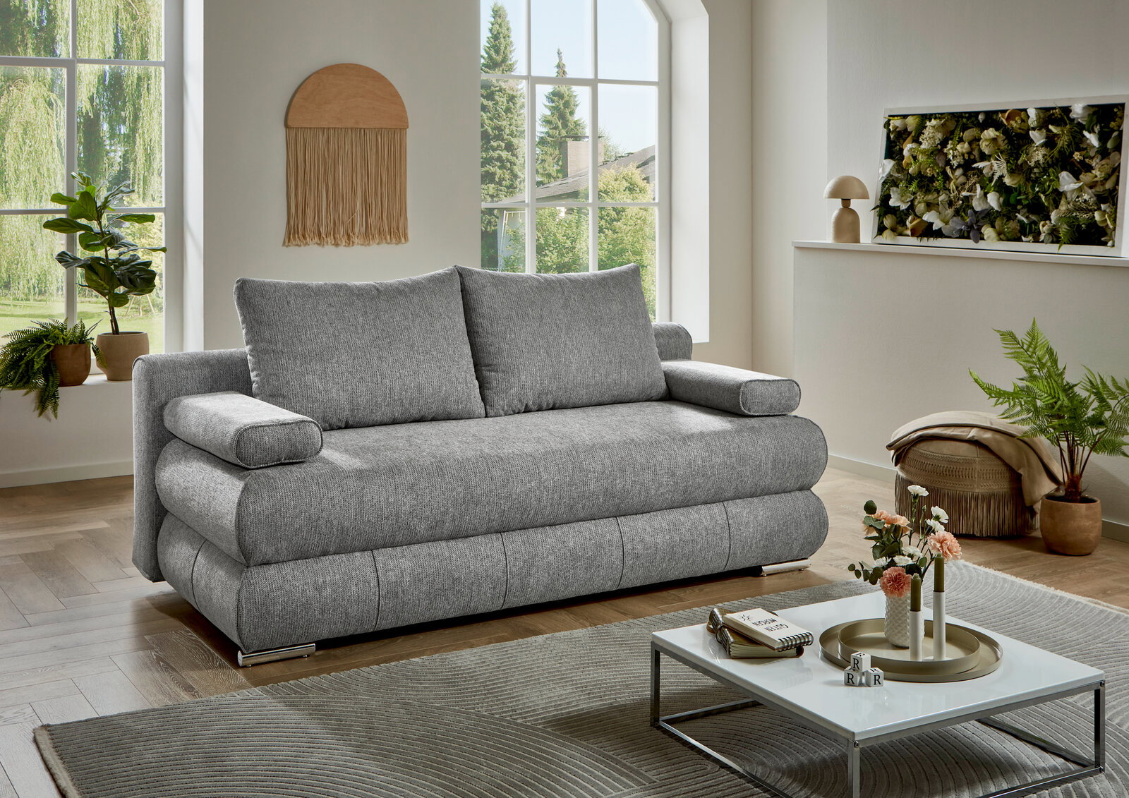 Graues Funktionssofa in einem modernen Wohnzimmer mit großen Fenstern und Pflanzen, seitliche Perspektive.