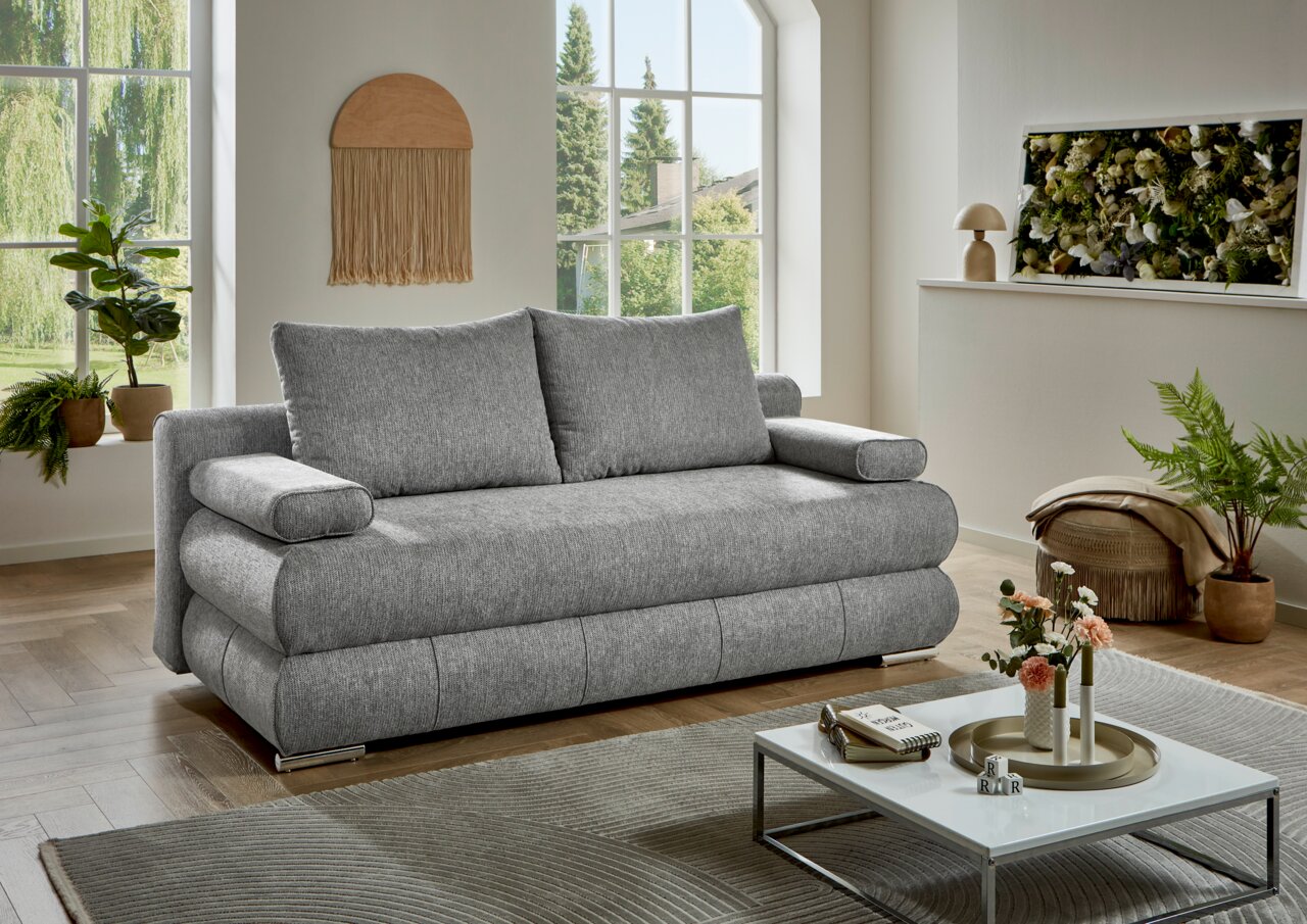 Graues Funktionssofa in einem modernen Wohnzimmer mit großen Fenstern und Pflanzen, seitliche Perspektive.