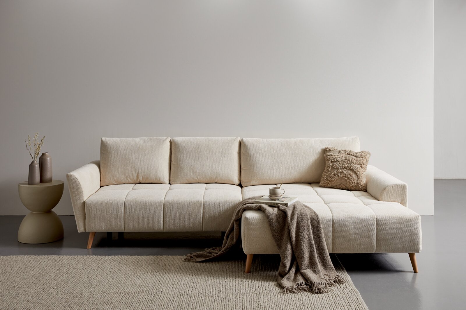 Z2 Ecksofa ZELDA Modernes beiges Ecksofa mit Chaise longue rechts, gestepptem Sitz und Holzfüßen, frontal aus leicht erhöhter Perspektive im minimalistischen Wohnzimmer mit Beistelltisch, Kissen und Decke.