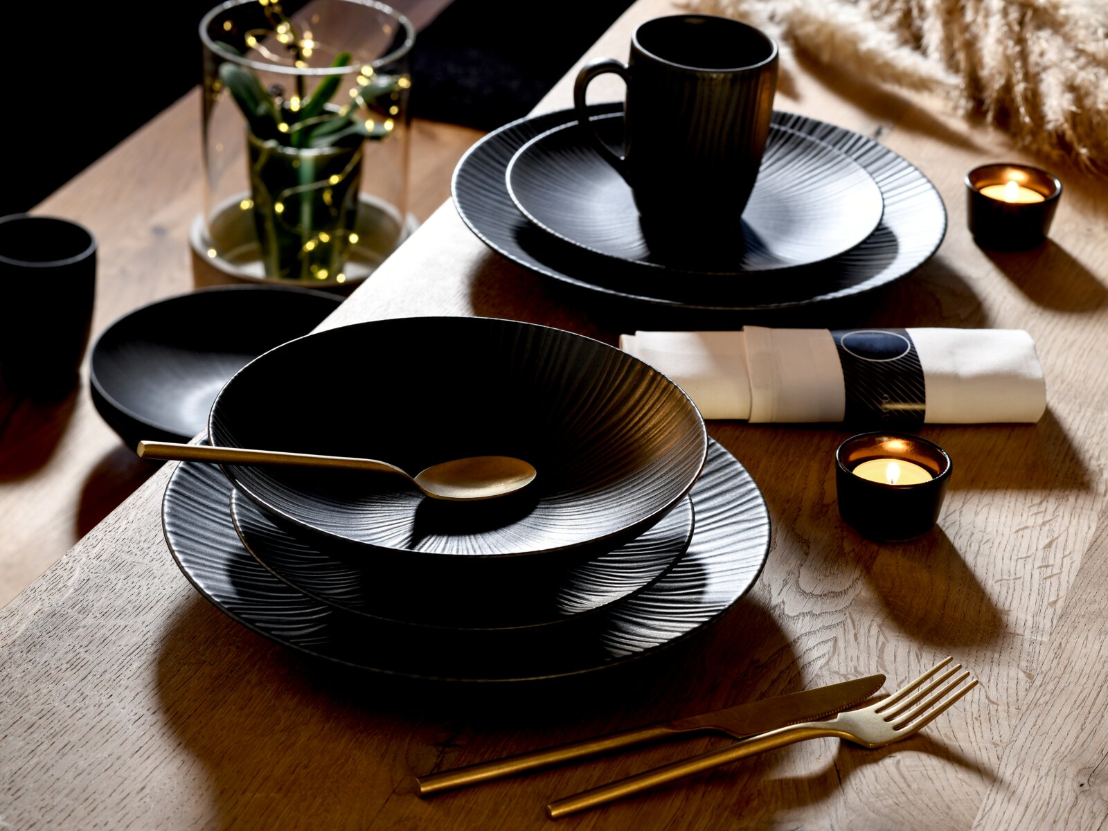 Schwarzes, geriffeltes Geschirr-Set mit tiefem Teller/Schale und flachen Tellern sowie schwarzer Tasse auf Holztisch, mit goldfarbenem Besteck, Serviette und Teelichtern Perspektive: leicht schräge Draufsicht.