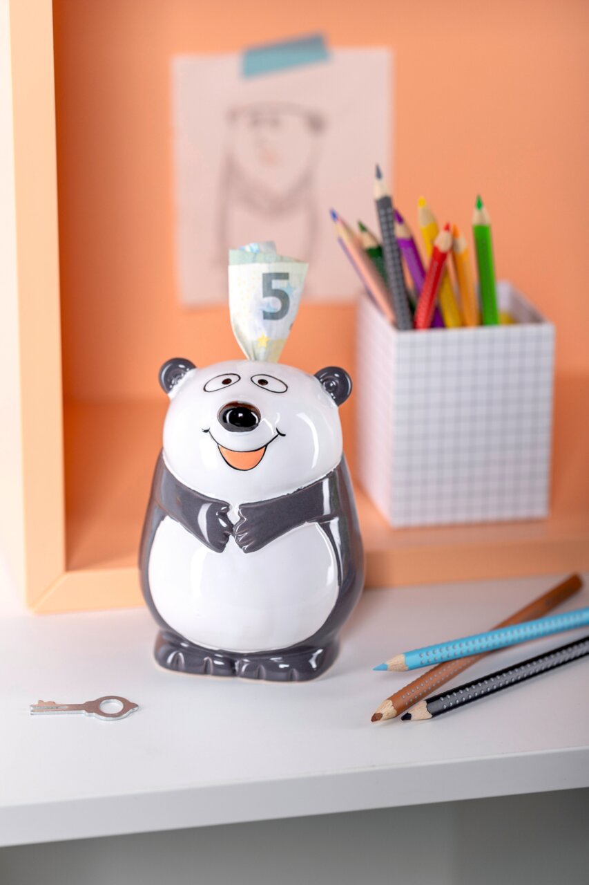 Vorderansicht einer niedlichen Panda-Spardose mit einem 5-Euro-Schein, umgeben von Buntstiften und einem Schlüssel auf einem Schreibtisch.