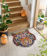 Bunte Fußmatte in Form eines Tigerkopfes mit abstraktem, geometrischem Design, platziert auf einem hellen Fliesenboden neben einer Holztreppe. Perspektive von oben.