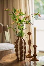 Zwei braune Kerzenständer mit eleganten Kurven, auf einem Tisch neben einer gestreiften Vase mit grünen Blättern und orangefarbenen Blüten, seitliche Perspektive.