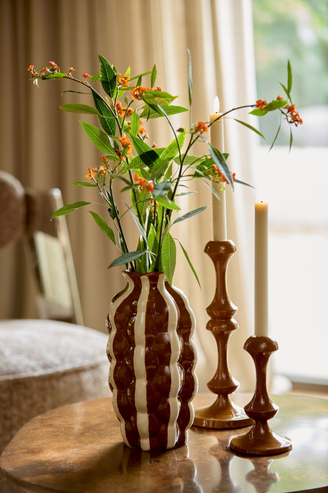 Zwei braune Kerzenständer mit eleganten Kurven, auf einem Tisch neben einer gestreiften Vase mit grünen Blättern und orangefarbenen Blüten, seitliche Perspektive.