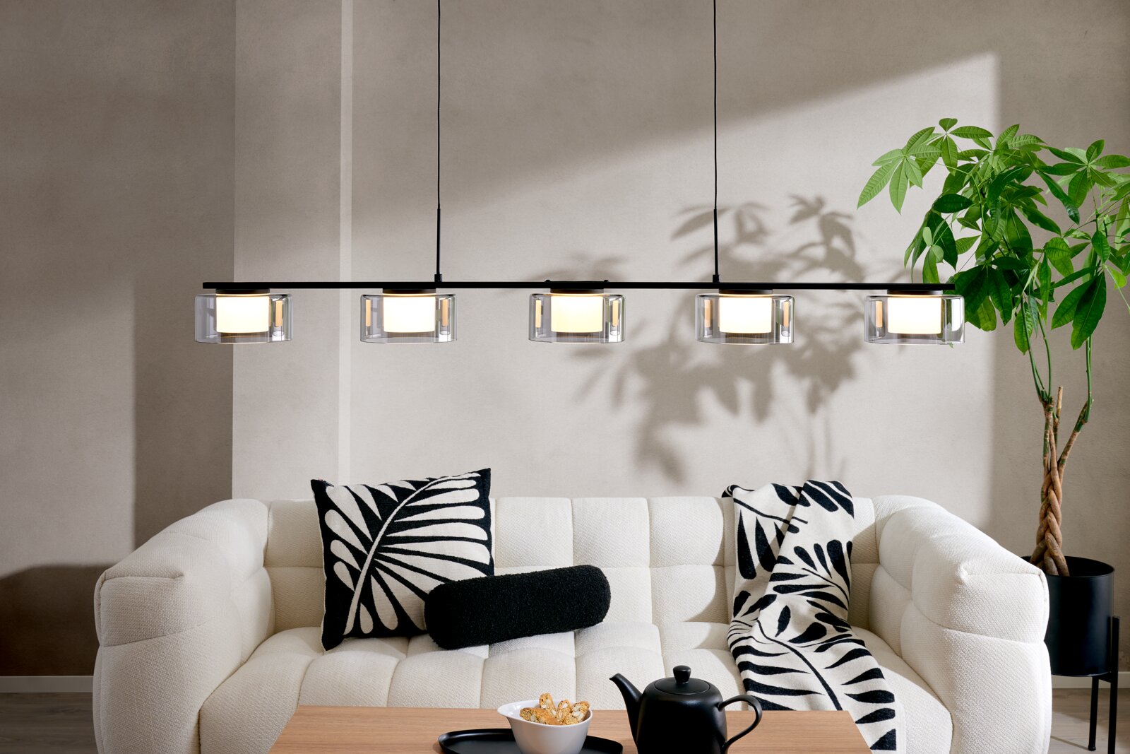 Moderne lineare Pendelleuchte mit fünf zylindrischen Glas-Schirmen an schwarzer Schiene über einem hellen Sofa im Wohnzimmer, Frontansicht