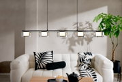 Moderne lineare Pendelleuchte mit fünf zylindrischen Glas-Schirmen an schwarzer Schiene über einem hellen Sofa im Wohnzimmer, Frontansicht
