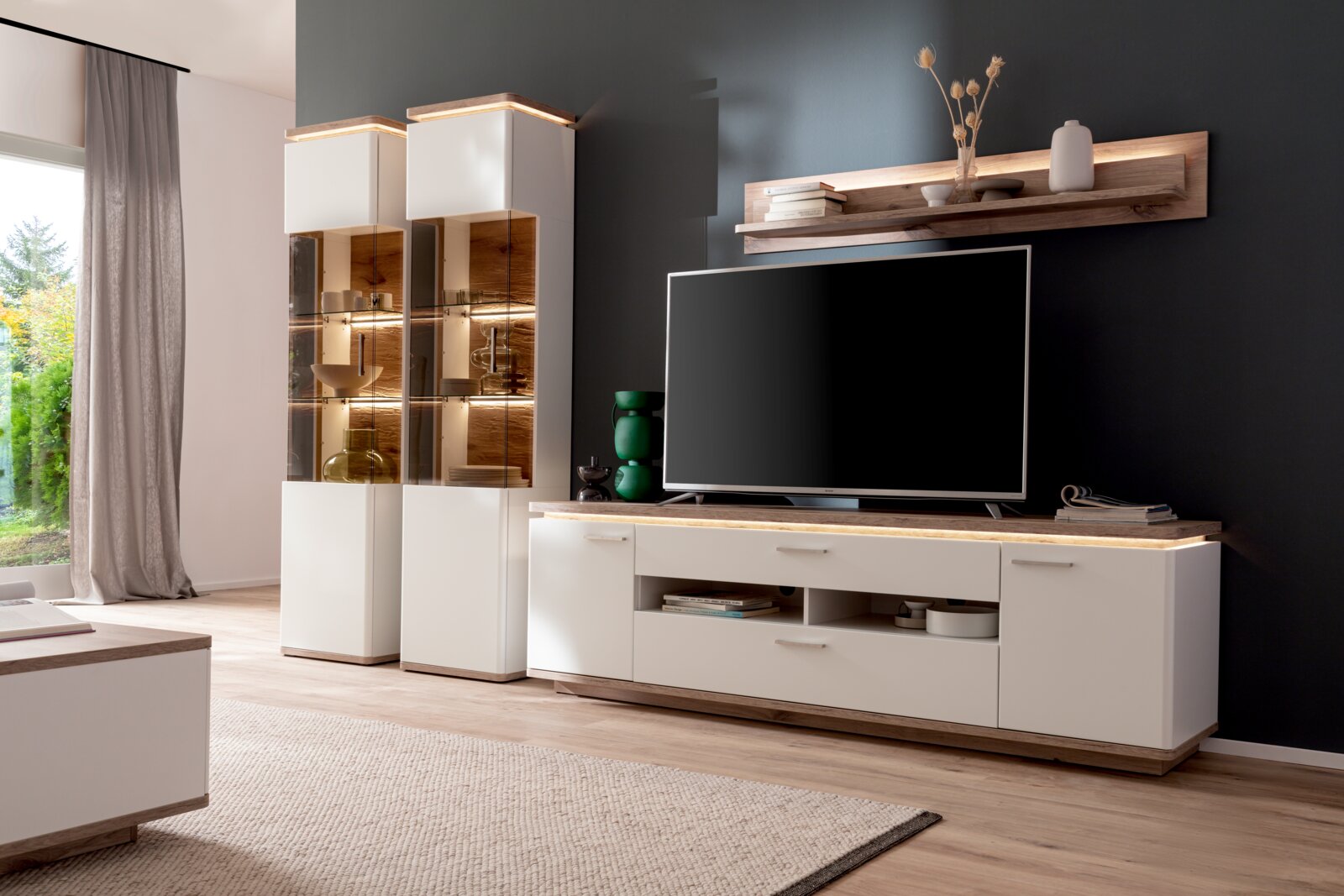 Modernes Wohnzimmermöbel-Set in Weiß mit Holzoptik: TV-Lowboard mit Schubladen, zwei beleuchtete Glasvitrinen und Wandboard über dem Fernseher, gezeigt in frontaler leicht schräger Perspektive.