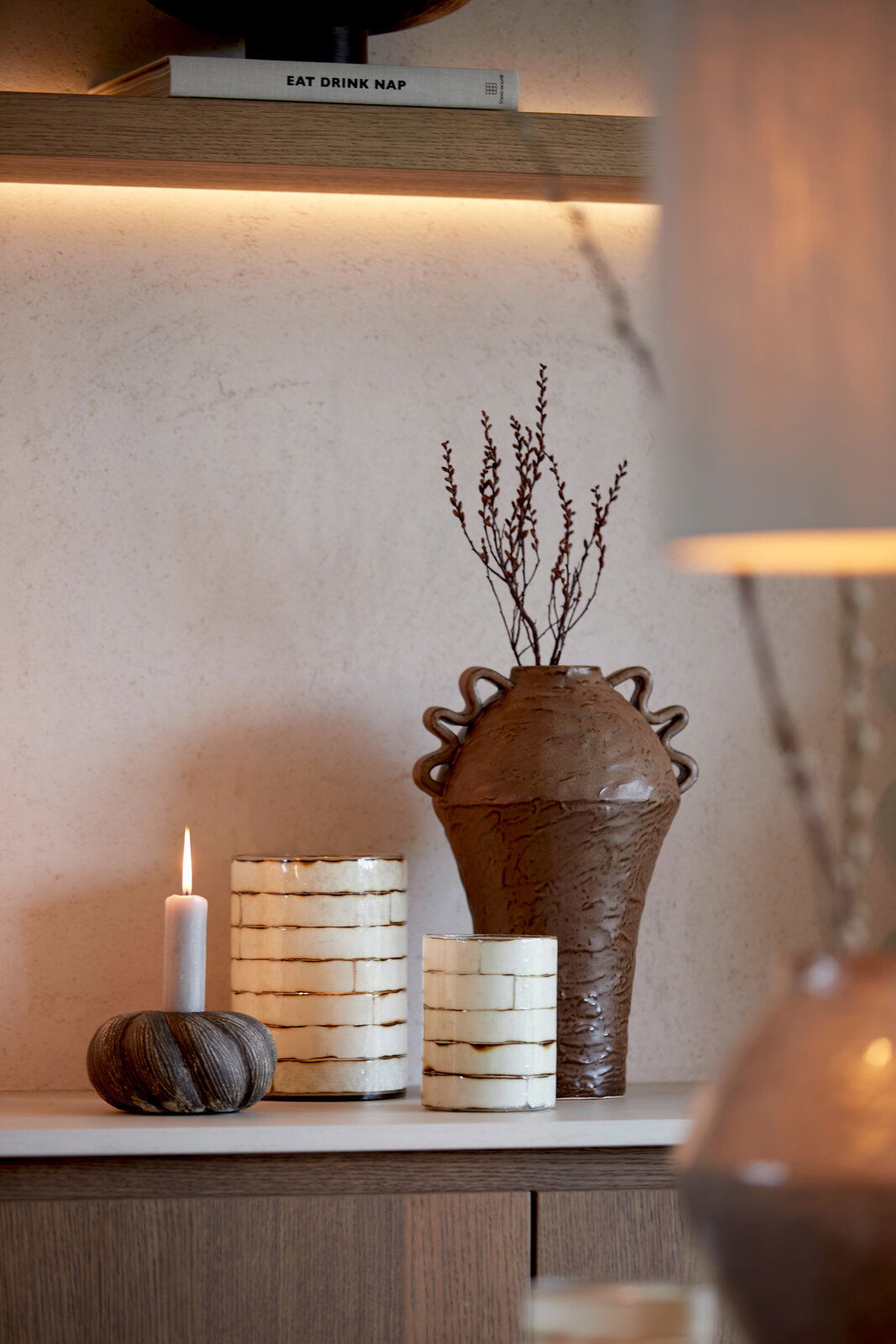 Light&Living Teelicht DRONERI 13cm Ein stilvolles Arrangement mit einem brennenden Teelicht in einem runden, geriffelten Halter, flankiert von zwei dekorativen Vasen und einem großen, kunstvoll gestalteten Krug. Die Perspektive zeigt die Objekte von vorne in einem gemütlichen Wohnambiente.