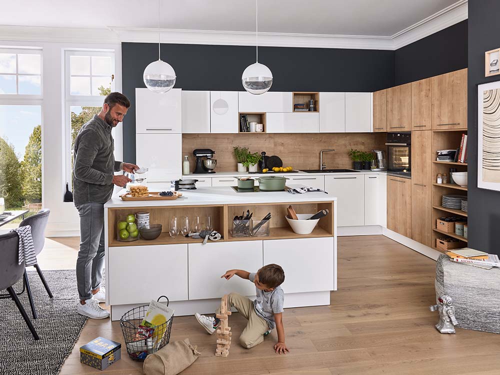 Moderne Küche in Weiß und Holzoptik, Kochinsel mit Regalen, spielendes Kind und Mann beim Zubereiten.