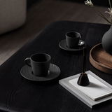 Schwarze Kaffeeuntertasse mit passender Tasse auf einem dunklen Holztisch, seitliche Perspektive.