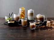 Verschiedene Getränke in doppelwandigen Gläsern, darunter Latte Macchiato, auf einem Tisch mit Kaffeebohnen und Zimtstangen, seitliche Perspektive.