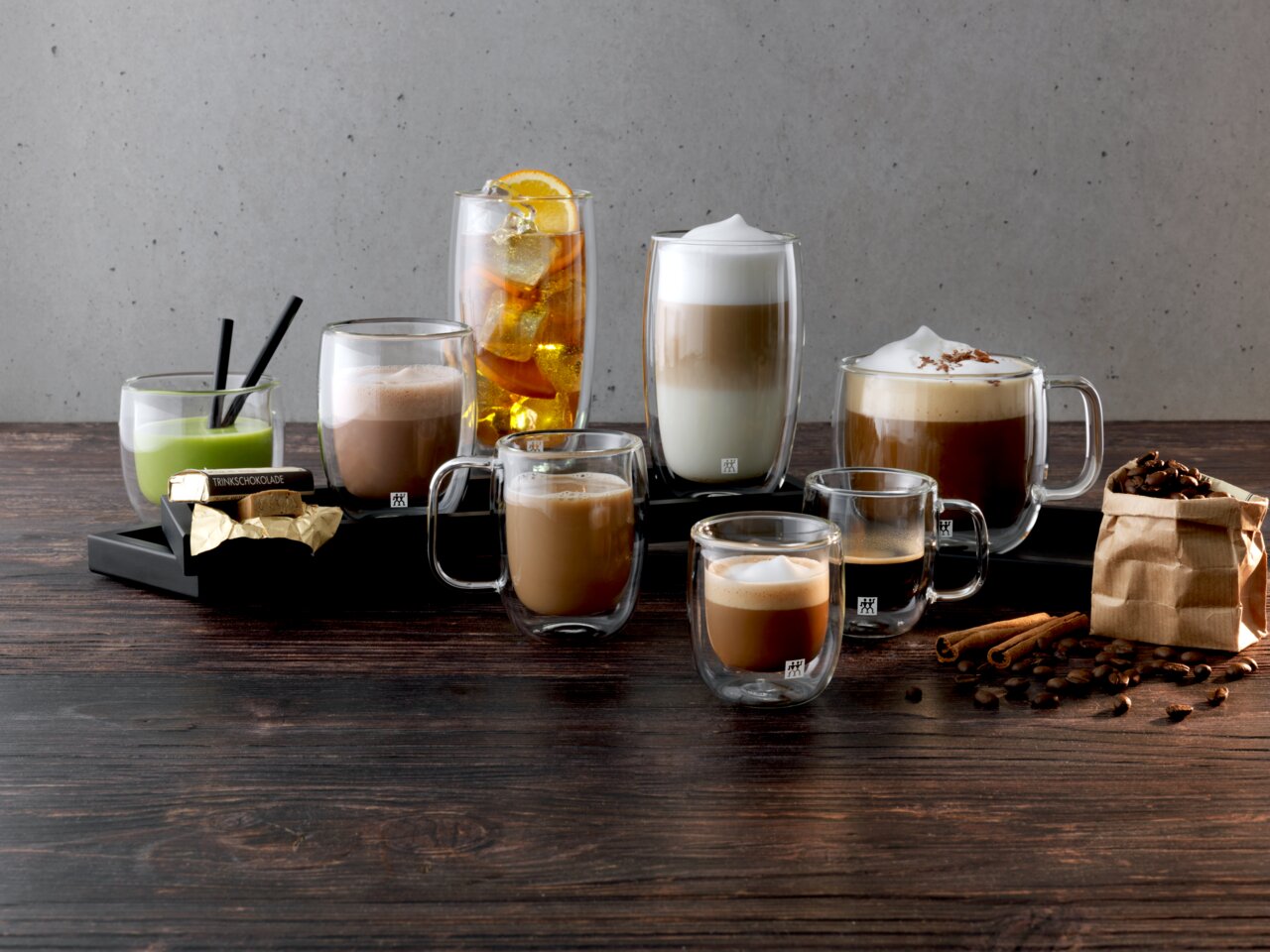 Verschiedene Getränke in doppelwandigen Gläsern, darunter Latte Macchiato, auf einem Tisch mit Kaffeebohnen und Zimtstangen, seitliche Perspektive.