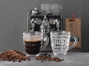 Zwei elegante, strukturierte Barista Kaffeebecher aus Glas, einer gefüllt mit Kaffee, auf einem Tisch mit Kaffeebohnen, im Hintergrund eine Kaffeemaschine. Frontalansicht.