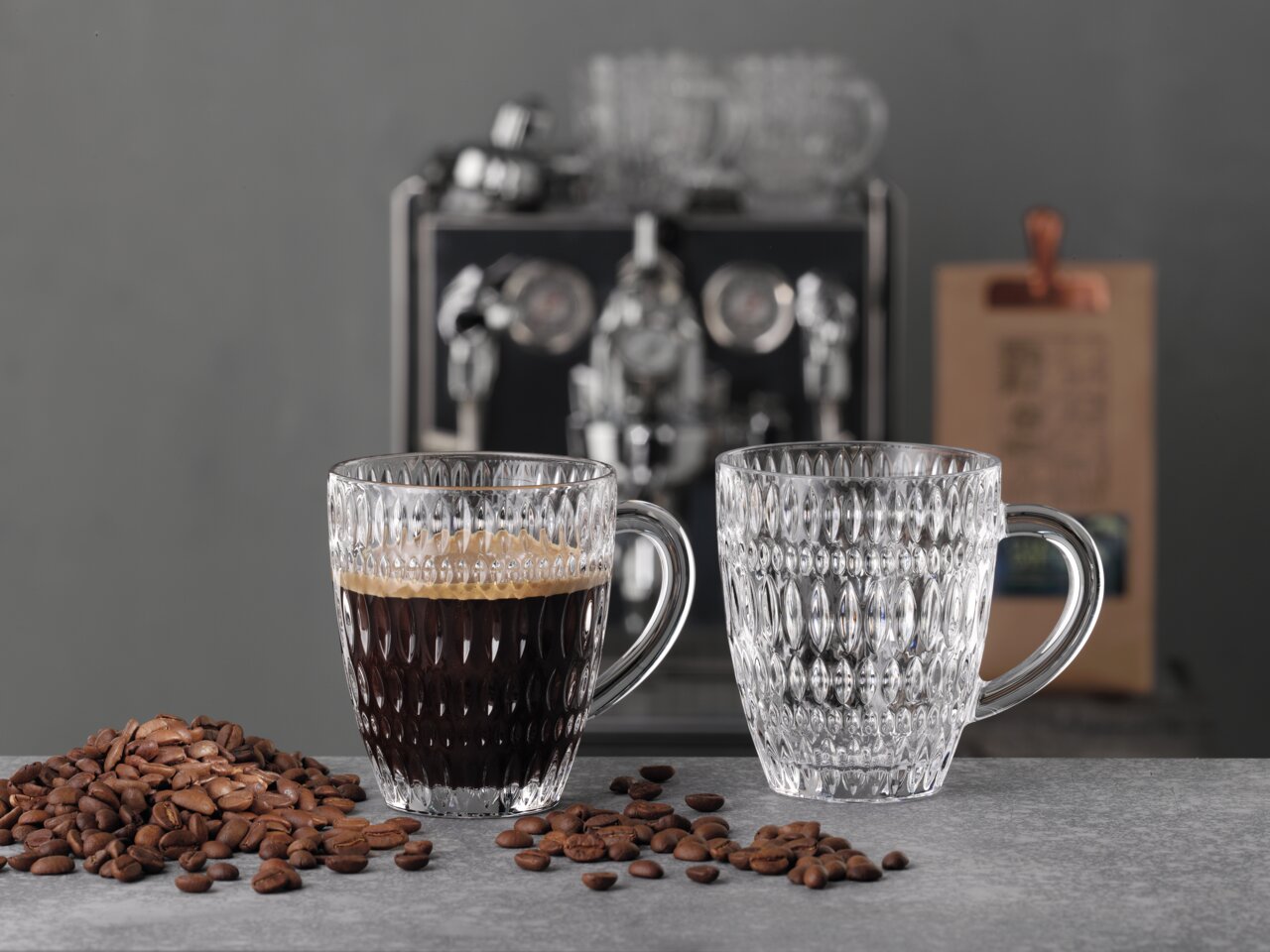 Zwei elegante, strukturierte Barista Kaffeebecher aus Glas, einer gefüllt mit Kaffee, auf einem Tisch mit Kaffeebohnen, im Hintergrund eine Kaffeemaschine. Frontalansicht.
