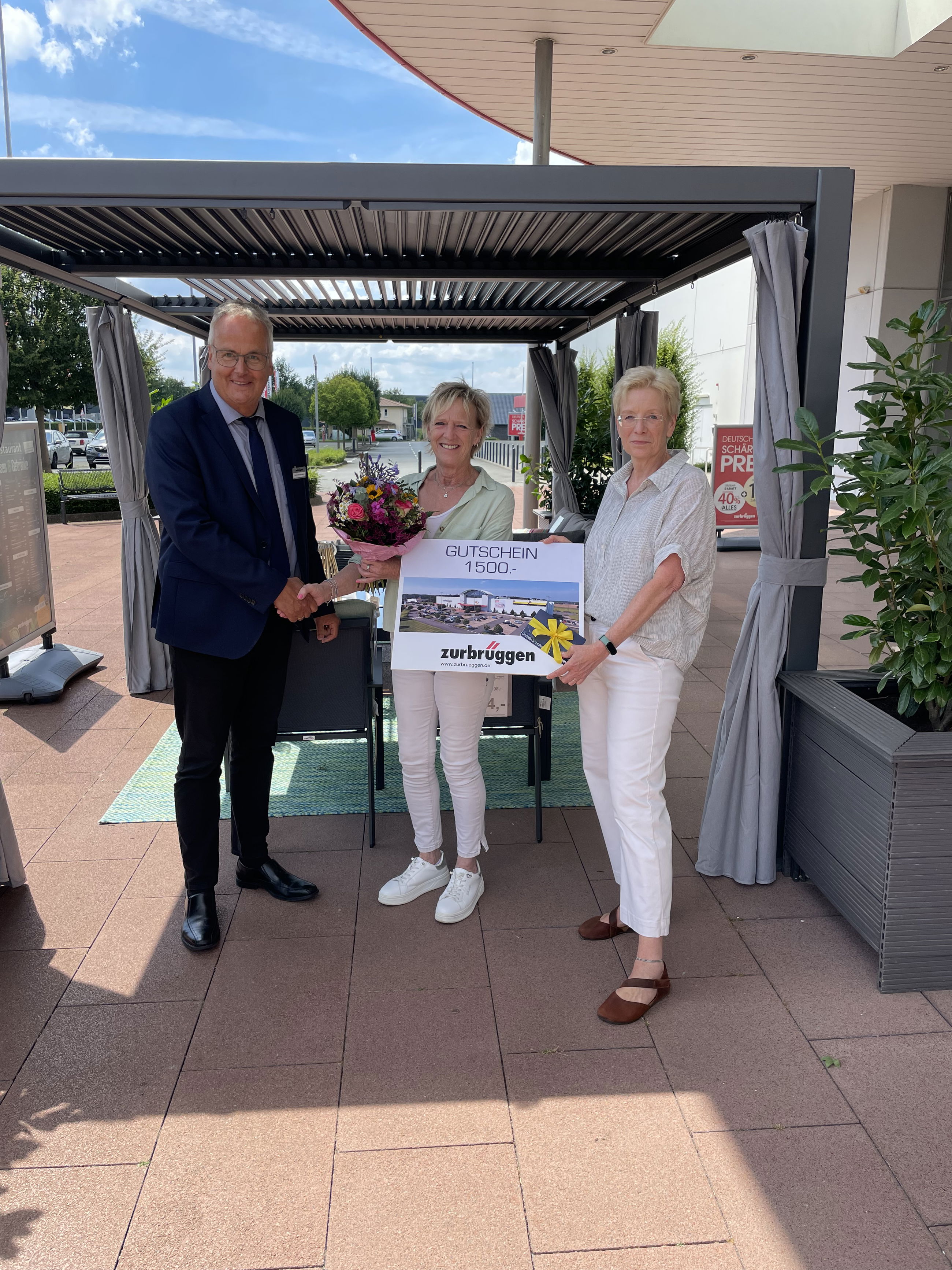 Das Bild zeigt drei Personen. Eine Frau hält einen Gutschein über „1.500 €“ von „Zurbrüggen“ in der Hand. Eine andere Frau hat einen Blumenstrauß. Ein Mann im Anzug schüttelt einer Frau die Hand. Die Übergabe findet draußen statt, unter einem Pavillon.