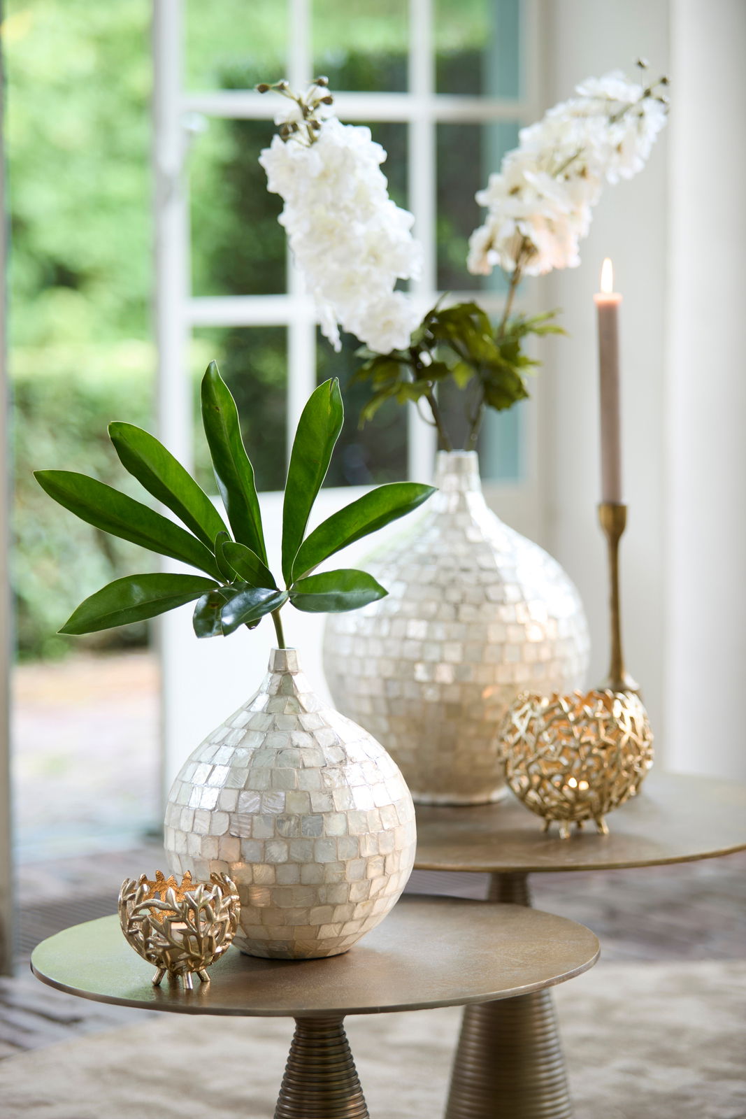 Elegantes Teelicht in goldener, filigraner Halterung auf einem Tisch, umgeben von dekorativen Vasen mit Blumen, aus seitlicher Perspektive fotografiert.