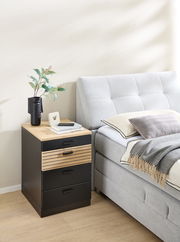 Rollcontainer mit drei schwarzen Schubladen und einer Holzschublade, neben einem grauen Bett, aus seitlicher Perspektive fotografiert.