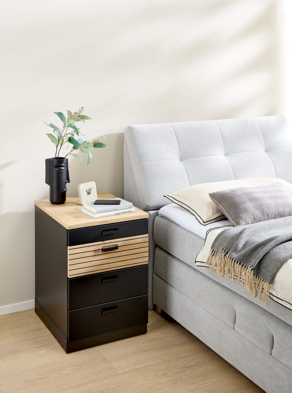 Rollcontainer mit drei schwarzen Schubladen und einer Holzschublade, neben einem grauen Bett, aus seitlicher Perspektive fotografiert.
