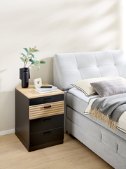 Rollcontainer mit drei schwarzen Schubladen und einer Holzschublade, neben einem grauen Bett, aus seitlicher Perspektive fotografiert.