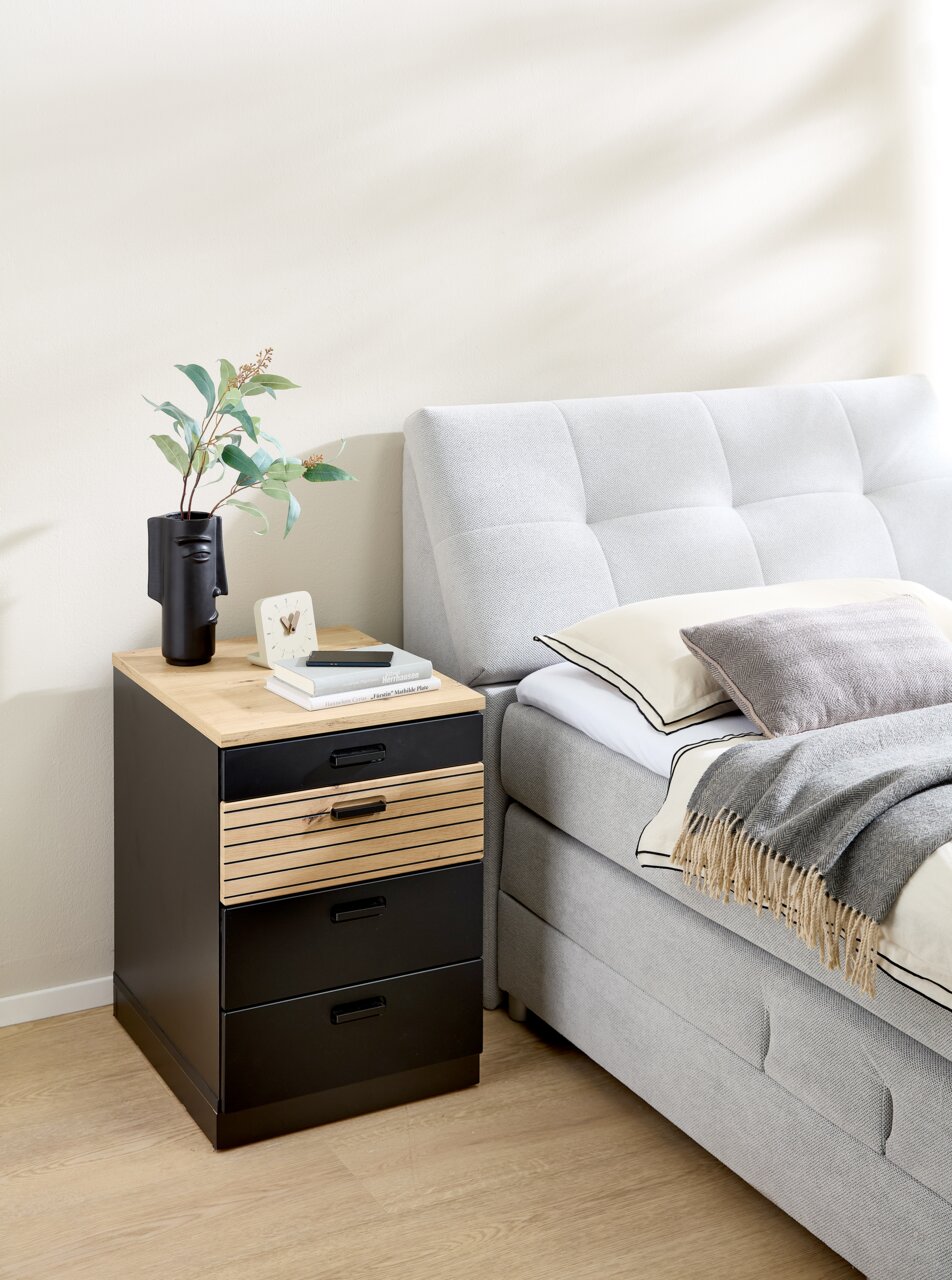 Rollcontainer mit drei schwarzen Schubladen und einer Holzschublade, neben einem grauen Bett, aus seitlicher Perspektive fotografiert.