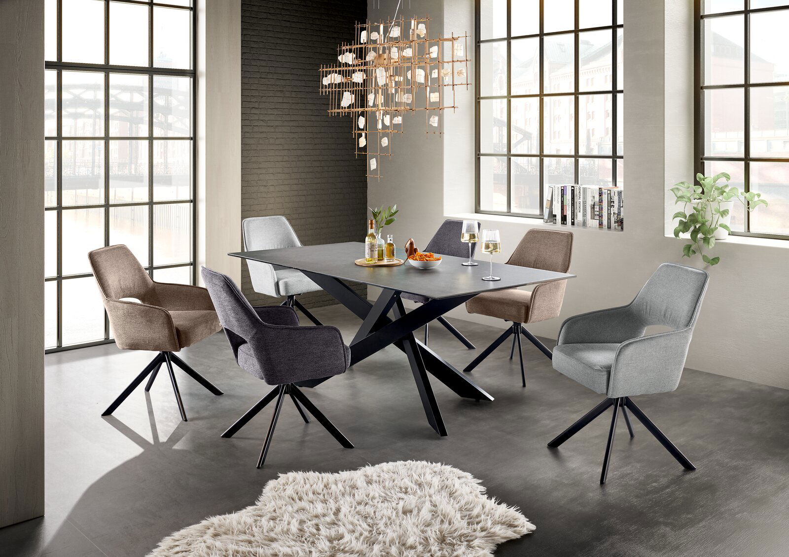 Set one by Musterring Esstisch BALTIMORE Moderner Esstisch mit schwarzer Tischplatte und X-förmigem Gestell, umgeben von sechs gepolsterten Stühlen in verschiedenen Farben, in einem hellen Raum mit großen Fenstern, aus der Perspektive von schräg vorne.