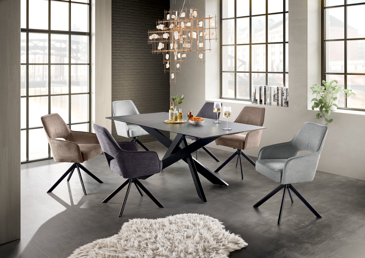 Moderner Esstisch mit schwarzer Tischplatte und X-förmigem Gestell, umgeben von sechs gepolsterten Stühlen in verschiedenen Farben, in einem hellen Raum mit großen Fenstern, aus der Perspektive von schräg vorne.