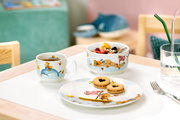 Kinder Geschirr-Set mit bunten Tiermotiven, bestehend aus Tasse, Schüssel und Teller, auf einem Tisch arrangiert. Perspektive von vorne.