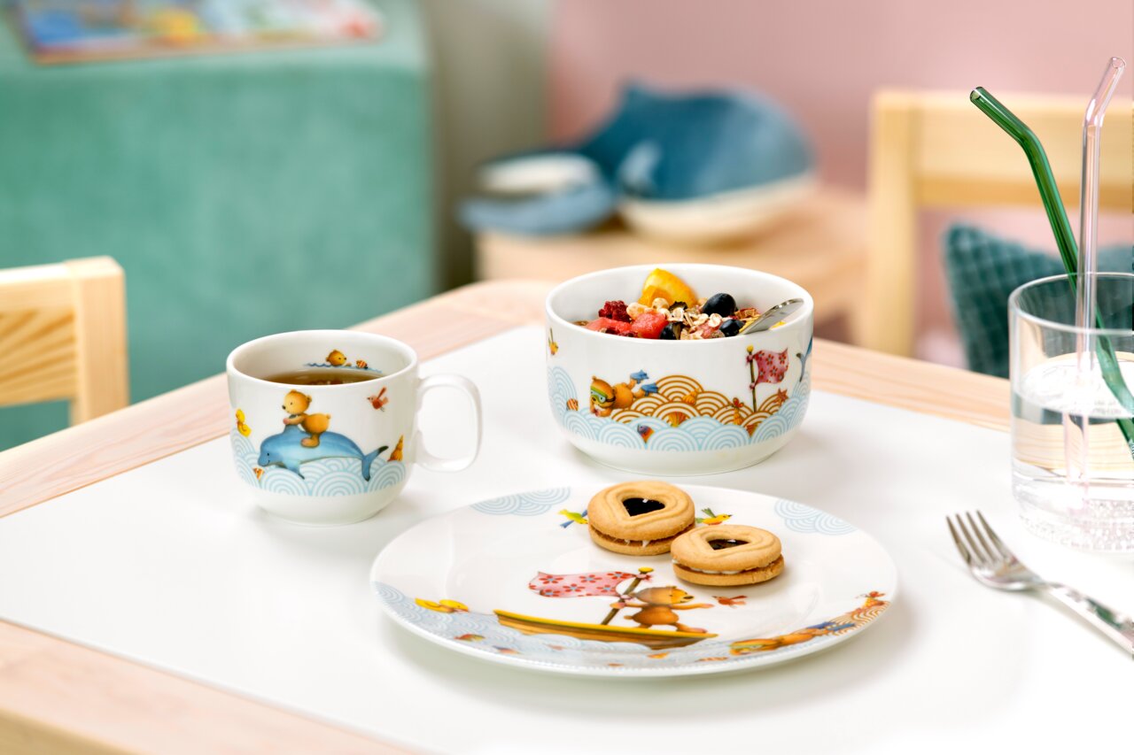 Kinder Geschirr-Set mit bunten Tiermotiven, bestehend aus Tasse, Schüssel und Teller, auf einem Tisch arrangiert. Perspektive von vorne.