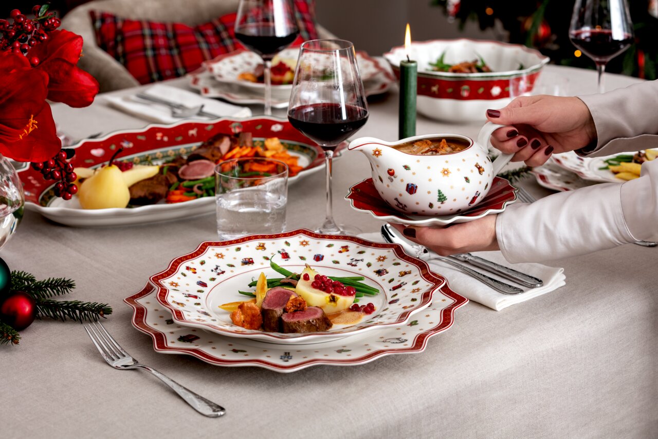 Festlich gedeckter Tisch mit weihnachtlichem Geschirr und Besteck, im Vordergrund ein Teller mit festlichem Essen und eine Hand, die eine Sauciere hält, seitliche Perspektive.