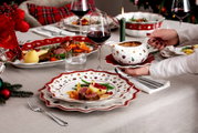 Festlich gedeckter Tisch mit weihnachtlichem Geschirr und Besteck, im Vordergrund ein Teller mit festlichem Essen und eine Hand, die eine Sauciere hält, seitliche Perspektive.
