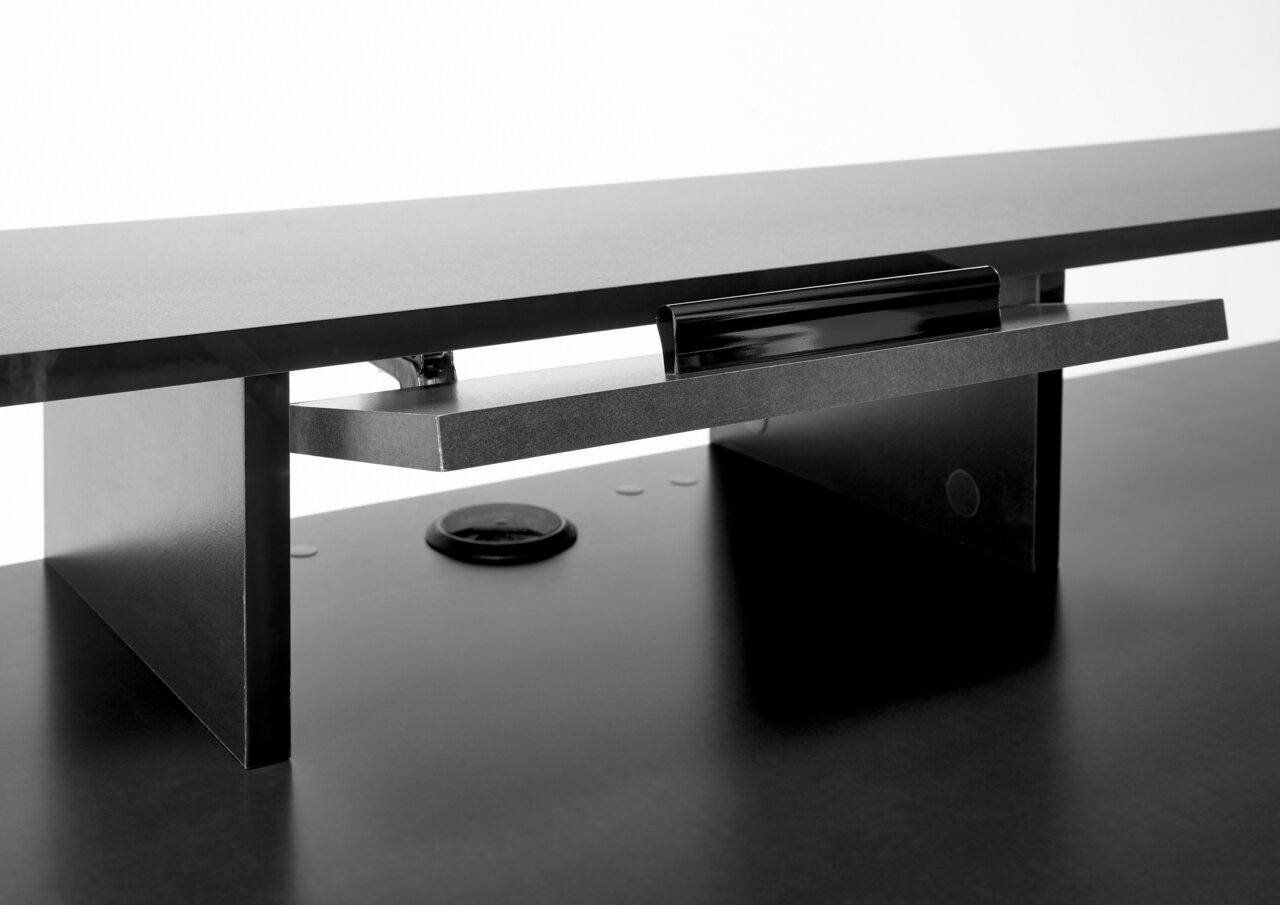 Nahaufnahme eines schwarzen Gaming-Schreibtisches mit erhöhtem Monitorständer und Kabeldurchführung, seitliche Perspektive.