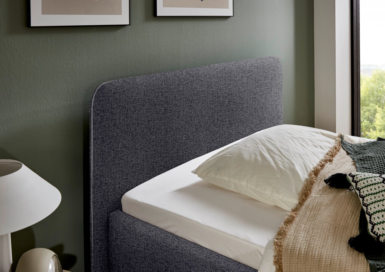 Seitliche Ansicht eines grauen Polsterbetts mit Kopfteil, dekoriert mit Kissen und Decke, neben einem Fenster.