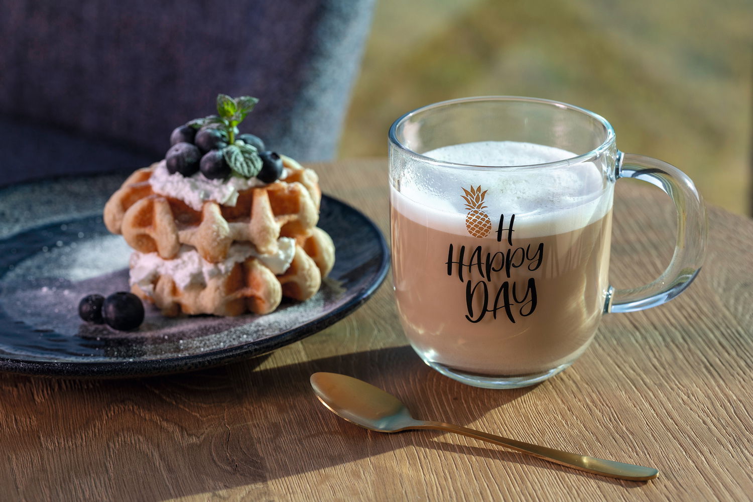 Leonardo Tasse EMOZIONE Glas-Tasse mit der Aufschrift 'Oh Happy Day' und einem Ananas-Design, gefüllt mit einem cremigen Getränk, auf einem Holztisch neben einem Teller mit Waffeln und Beeren, seitliche Perspektive.