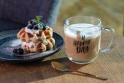 Glas-Tasse mit der Aufschrift 'Oh Happy Day' und einem Ananas-Design, gefüllt mit einem cremigen Getränk, auf einem Holztisch neben einem Teller mit Waffeln und Beeren, seitliche Perspektive.